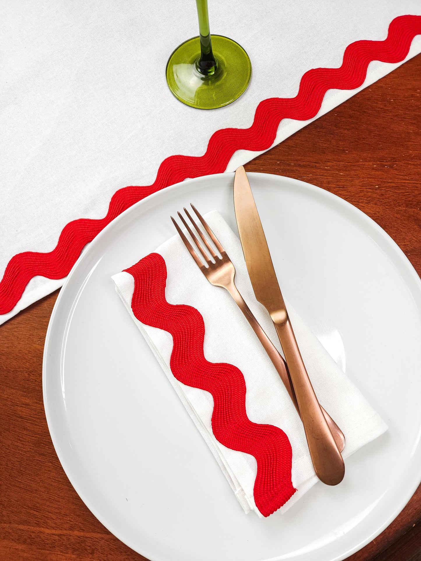 A festive cloth napkin made of white cotton with extra wide ric rac trim on one edge shown on a plate with copper silverware.